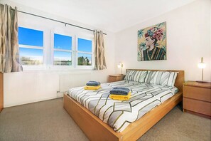 Un dormitorio con cama, ventana, mesita de noche, lámpara y un cuadro en la pared.