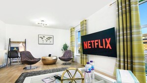 Un salon moderne doté d’un grand téléviseur à écran plat affichant le logo « NETFLIX », de deux fauteuils en cuir, d’une table basse ronde, d’un globe terrestre et d’une bibliothèque.