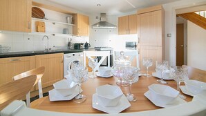 Une cuisine moderne avec des meubles en bois, une table à manger dressée pour six personnes, avec de l’argenterie blanche et des verres en cristal, et un four encastré.