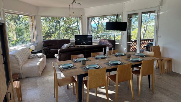 A dining area with a wooden table set for four, surrounded by wooden chairs. A brown leather sofa and a white couch are positioned against the wall, with a flat-screen TV mounted above the sofa. Large windows provide natural light and a view of greenery outside. - Rental Villa, 200m², 6 bedrooms, sleeps up to 12, swimming pool, Montauroux (Montauroux)