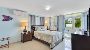 A bedroom with a bed, bedside lamps, a nightstand, a wooden bench, an air conditioner, and a window with curtains. - Steps to Beach/Pool & Jacuzzi/Wave Coast Suite 8! (Rincon)