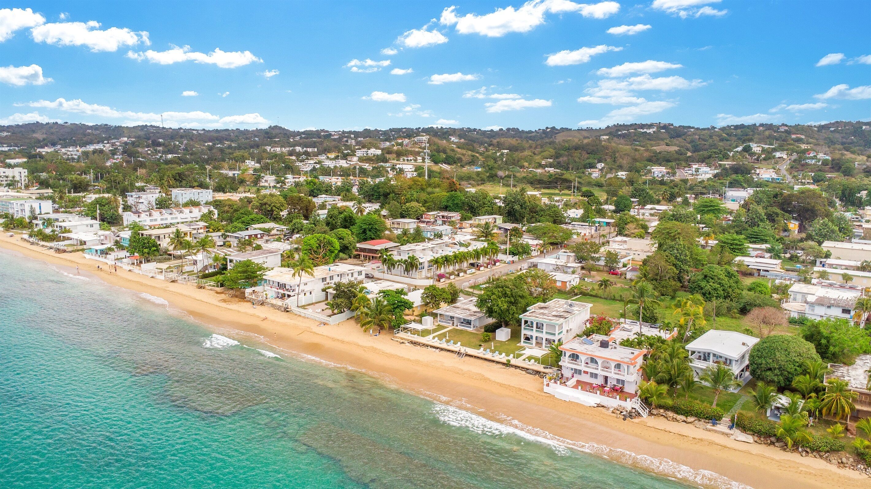 A coastal town with beachfront properties, clear skies, and a hilly backdrop.