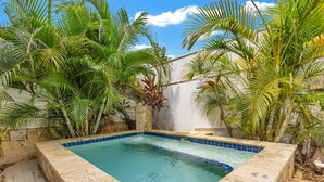 A rectangular pool with a stone border, surrounded by lush green palm trees and a white wall. - Steps to Beach/Pool & Jacuzzi/Wave Coast Suite 8! (Rincon)