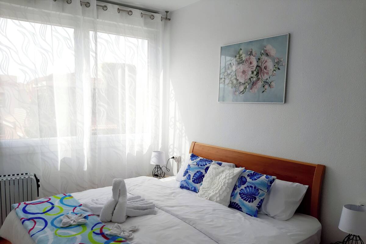 Een net opgemaakt bed met witte lakens, een kleurrijk dekbed en een zachte witte handdoek. Er is een wandlamp en een schilderij van bloemen.