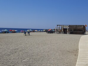 Uma praia com guarda-sóis e cadeiras de praia, uma área de bar e uma vista clara do oceano.