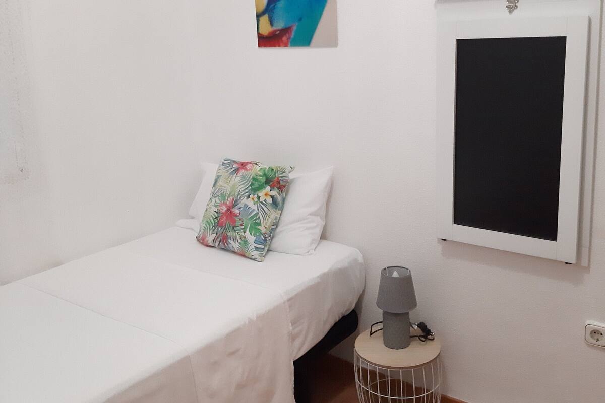Een slaapkamer met een enkel bed, een kleurrijk kussen, een aan de muur hangend kunstwerk, een klein tafeltje met een lamp, en een deur met een zwart scherm.