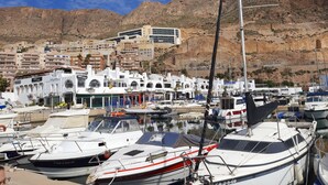 Un puerto deportivo con barcos amarrados, un edificio con una bandera y montañas al fondo.