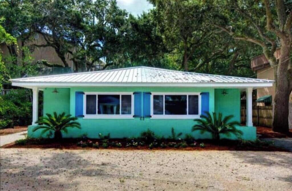 A small, single-story house with a green exterior and blue trim, surrounded by trees and a gravel driveway.