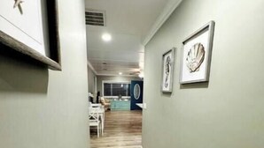 A hallway with a wooden floor, framed artwork on the walls, and a view into a room with a blue accent wall and a round mirror. - ♥ of SSI Bungalow, Quick walk to beach and food ☼ (St. Simons Island)