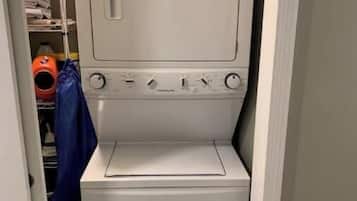 Una lavadora y secadora de carga frontal, de color blanco y tamaño compacto, en un cuarto de lavado con un estante pequeño en la parte superior.