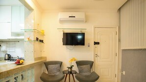A modern living room with a TV, two grey armchairs, a small table with flowers, and a kitchen area with a fruit bowl. - 360 Aterro do Flamengo By SUHCasa (Rio de Janeiro)