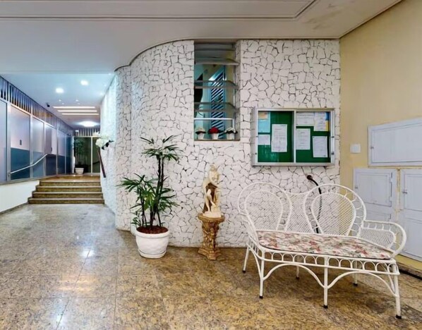 Un pasillo con una pared blanca con textura, una estatua, una planta en maceta y una silla metálica blanca.