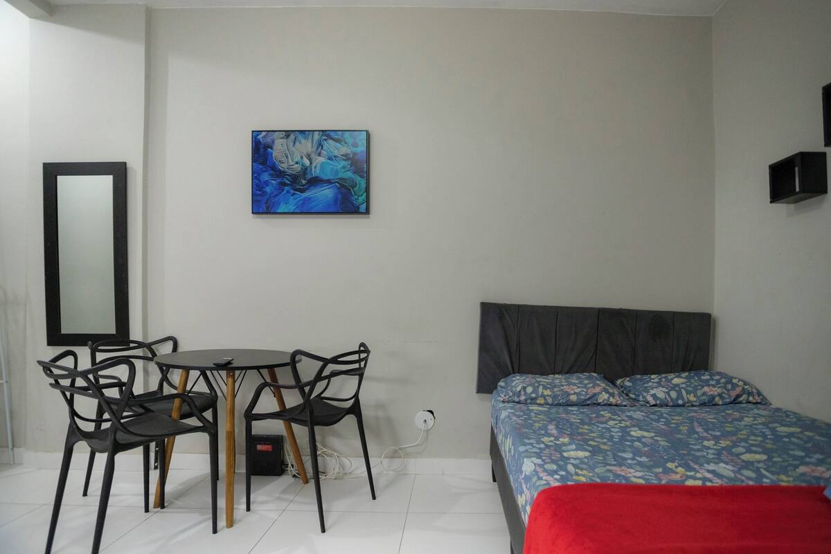 Un dormitorio con una cama, una mesita de noche, una silla, un escritorio con silla, un cuadro en la pared y un espejo.
