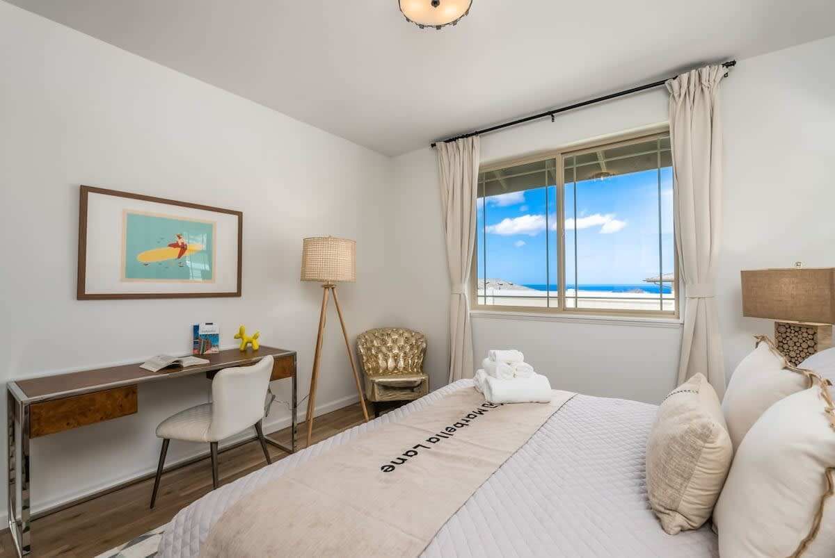 A hotel room with a bed, a desk, a chair, a lamp, and a window with a view of the sea.