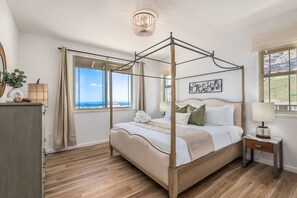 Una cama con dosel, una mesita de noche con lámpara, vista al mar a través de amplias ventanas y suelo de madera.