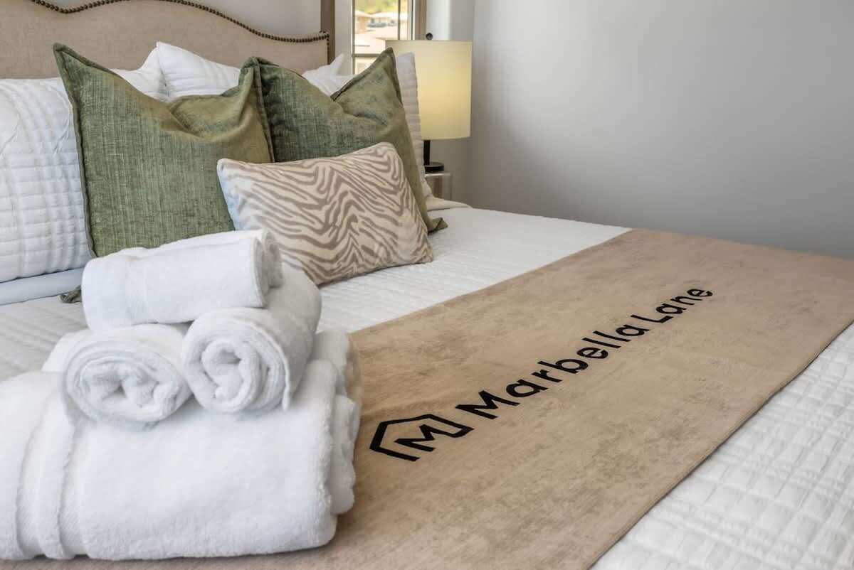 A neatly made bed with rolled white towels, a beige throw blanket with a logo, and green and white pillows.