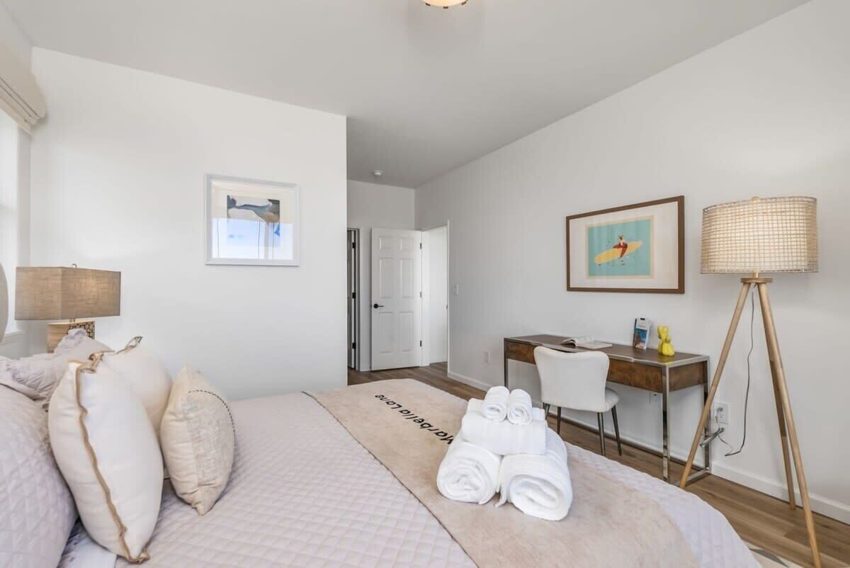 A hotel room with a bed, a desk, a chair, a lamp, and a door.