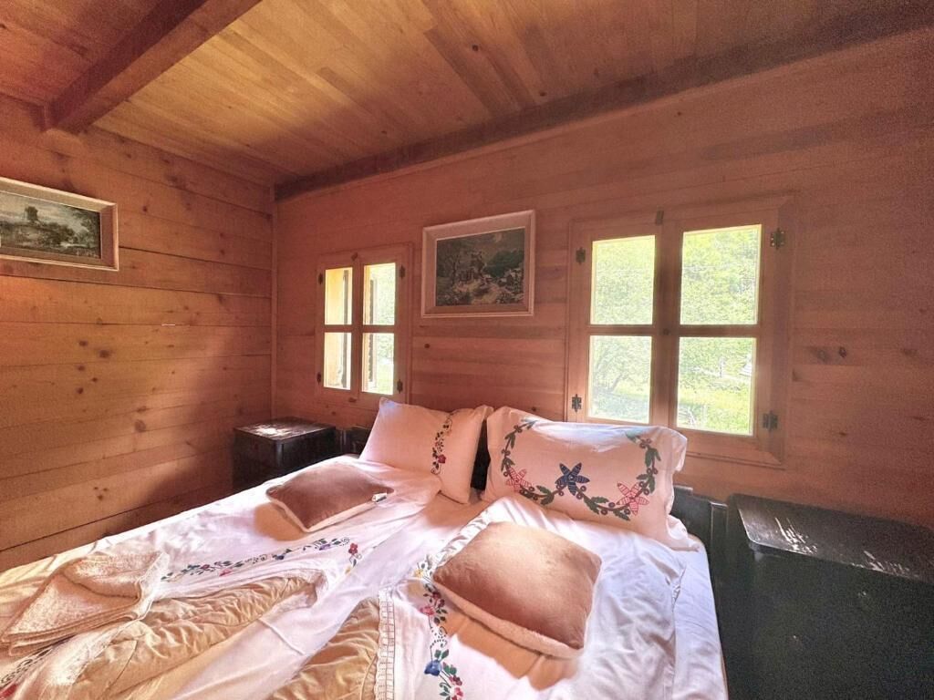 A wooden cabin room with a bed, two windows, and a painting on the wall.