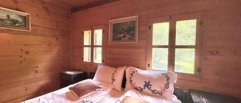 A wooden cabin room with a bed, two windows, and a painting on the wall.