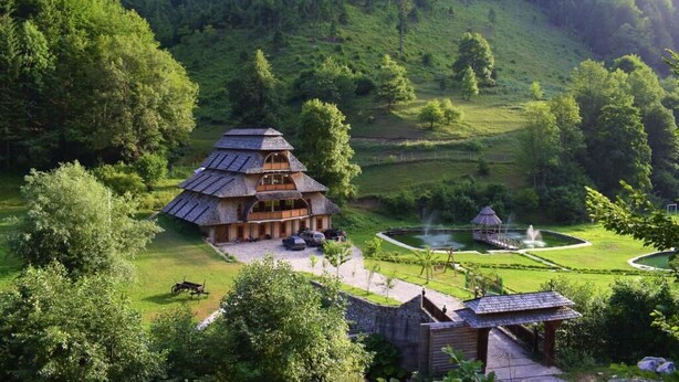 Une maison traditionnelle en bois, dotée d’un toit à pente, entourée d’une végétation luxuriante et d’un petit étang agrémenté d’une fontaine.