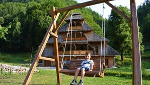 Un ensemble de balançoires en bois avec un enfant assis dessus, une maison en bois rustique en arrière-plan et une zone herbeuse avec des arbres.