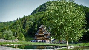 Un chalet traditionnel en bois, doté d’un toit à pente, situé au bord d’un étang paisible, entouré d’une végétation luxuriante et d’une colline boisée.