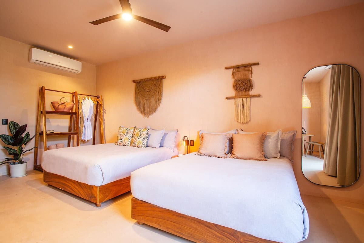 A hotel room with two beds, a ceiling fan, an air conditioning unit, a mirror, and a small table with a chair.