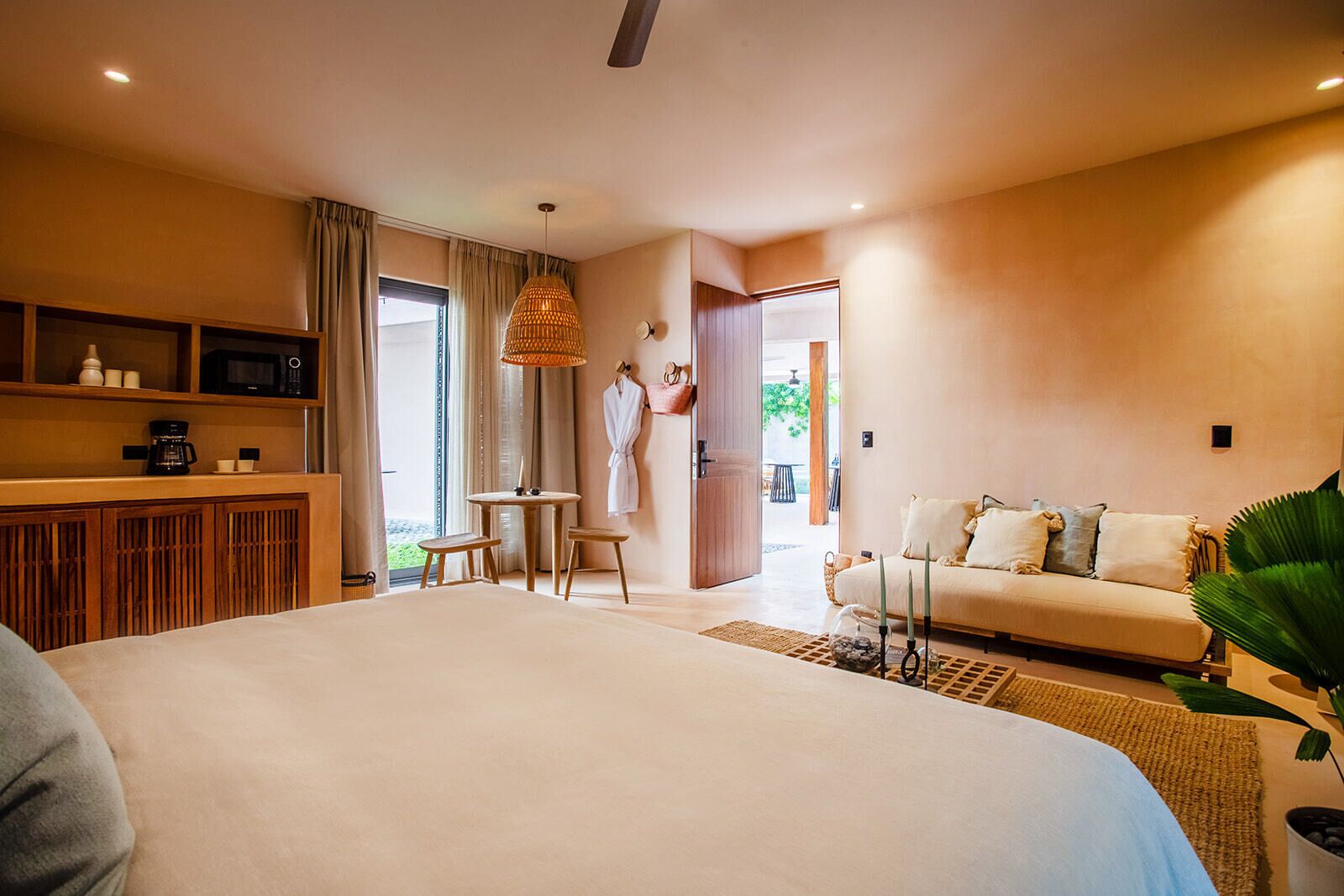 A modern hotel room with a large bed, a sitting area with a table and chairs, a sofa, and a view of the outdoors.