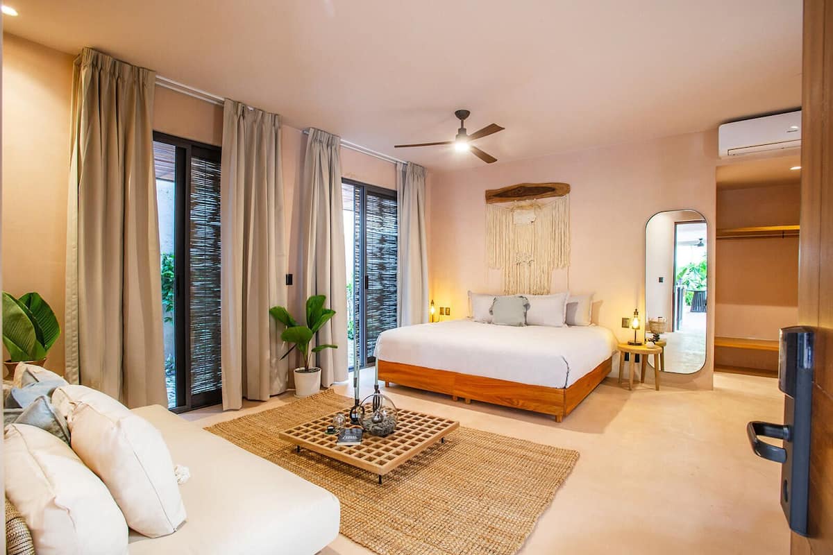 A bedroom with a bed, a ceiling fan, a large mirror, a bedside table with a lamp, and a view of the outdoors through the window.