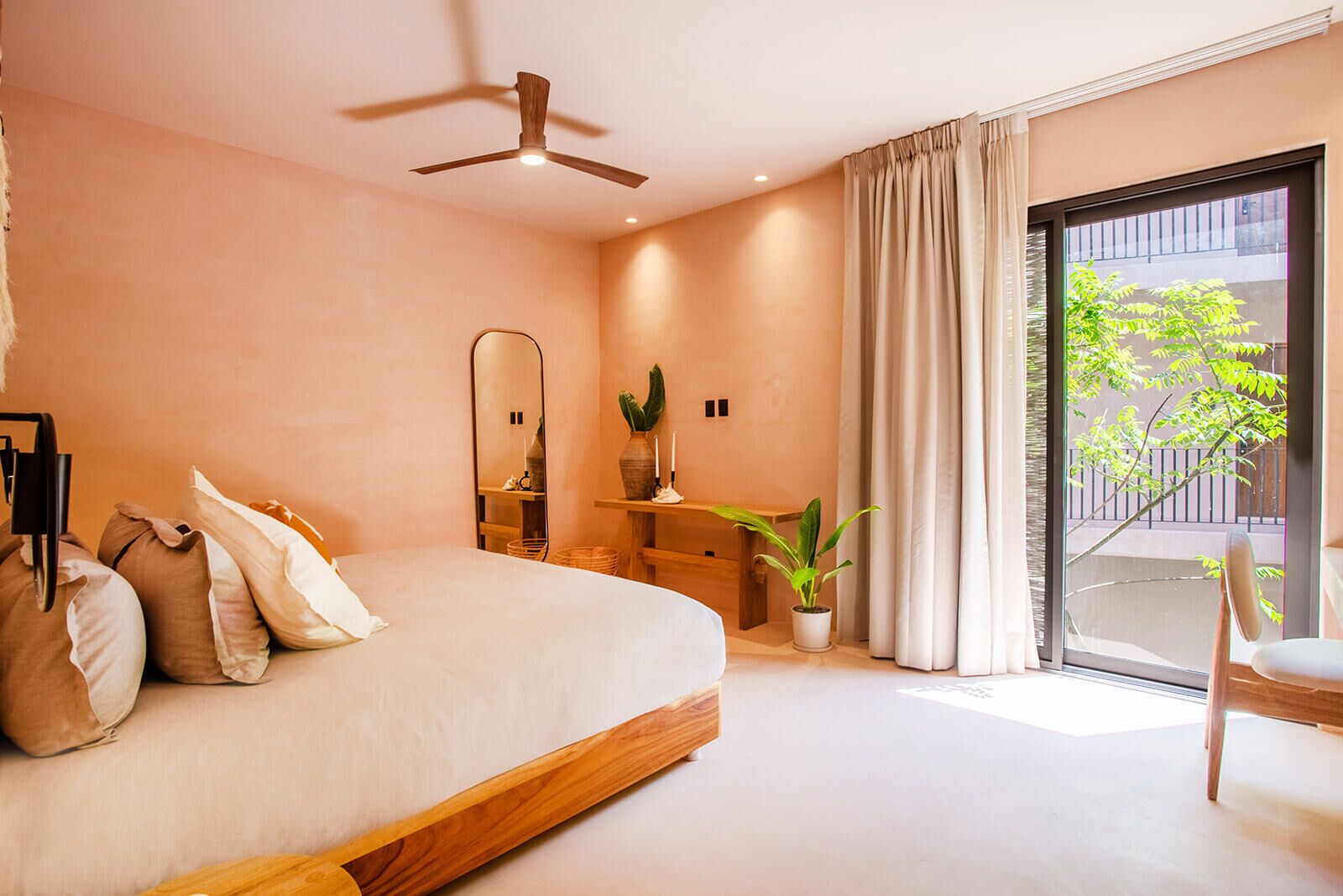 A modern bedroom with a large bed, a ceiling fan, a bedside table with a mirror, a potted plant, and a view of the outdoors through a sliding door.