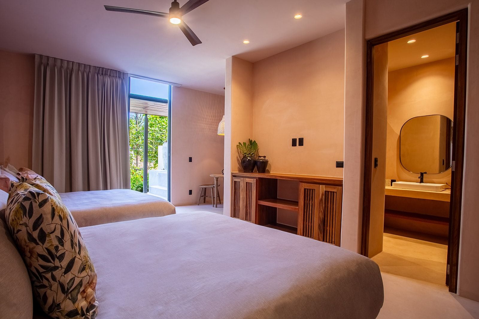 A modern hotel room with two beds, a desk with a mirror, and a view of greenery outside.