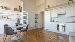 A modern kitchen with white cabinets, a built-in dishwasher, and a herringbone wood floor. There is a dining area with a round table and chairs, and shelves with plants and decorative items.