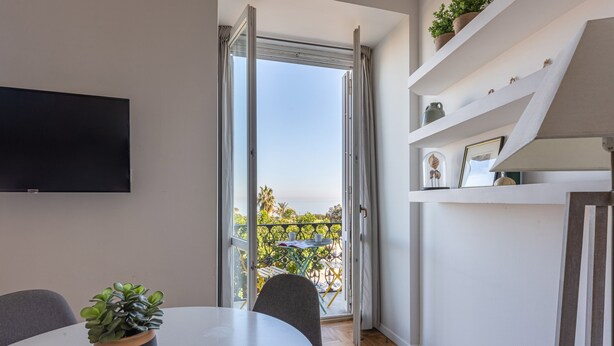 A modern living room with a flat-screen TV, a dining table with chairs, a potted plant, and a staircase with shelves.