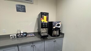 Un espace dédié au café, avec un panneau indiquant les heures du petit-déjeuner, une machine à café, un distributeur de café et un comptoir où l'on trouve des tasses et un menu.