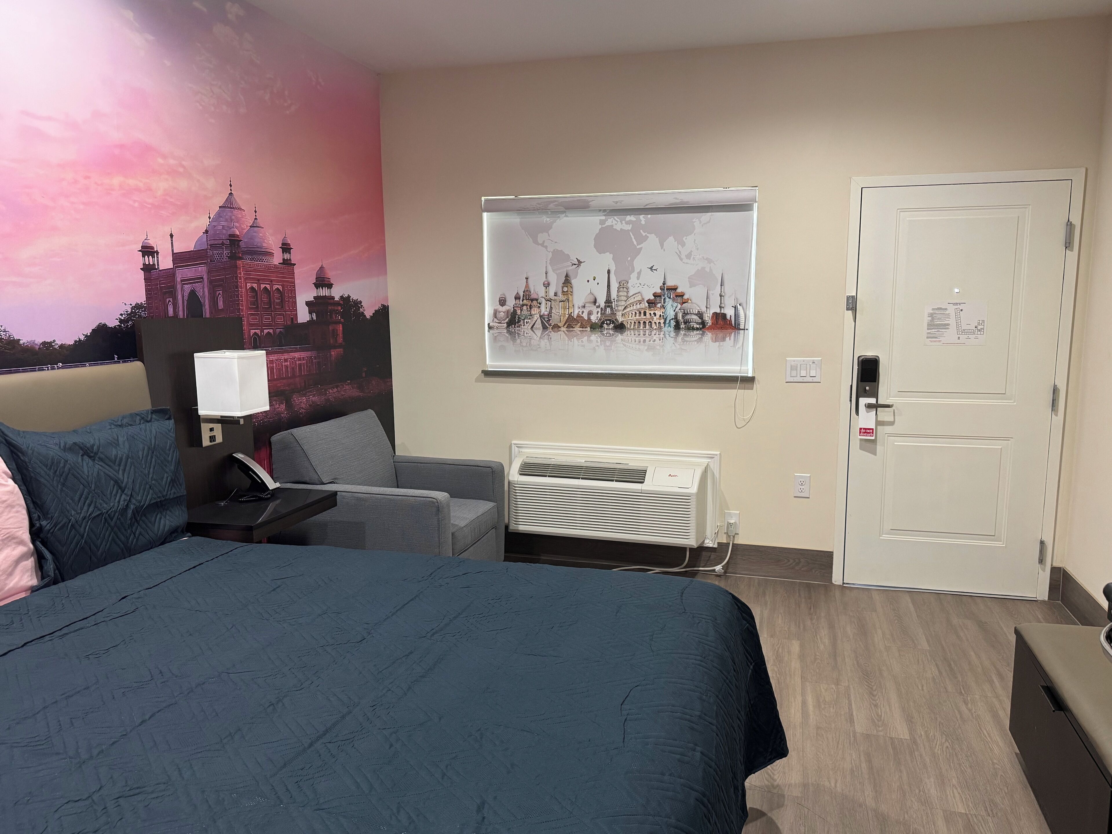 Habitación de hotel con cama, escritorio, silla, aire acondicionado instalado en la pared y una puerta con cerradura de llave electrónica.