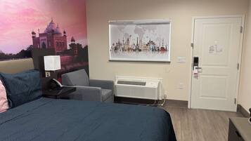 A hotel room with a bed, a desk, a chair, a wall-mounted air conditioner, and a door with a keycard lock.