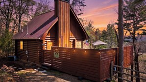 A wooden cabin with a chimney, a porch with string lights, and a flag.