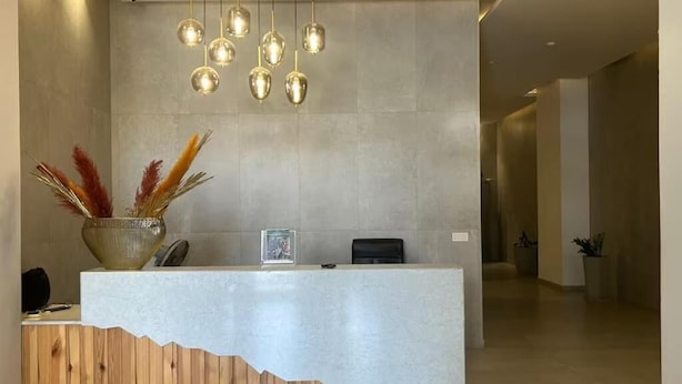 A reception desk with a wooden base and a white top, a vase with dried pampas grass, and a hanging light fixture with multiple bulbs. - Waypoint Suites (Dakar)