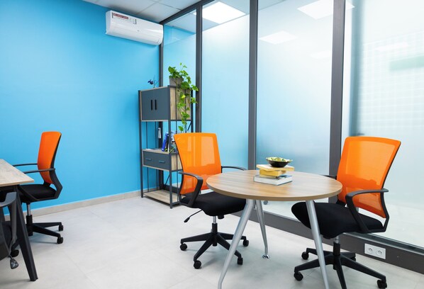Une salle de conférence avec une table ronde, quatre chaises de bureau orange, une petite plante et un mur bleu sur lequel est installé un appareil de climatisation.
