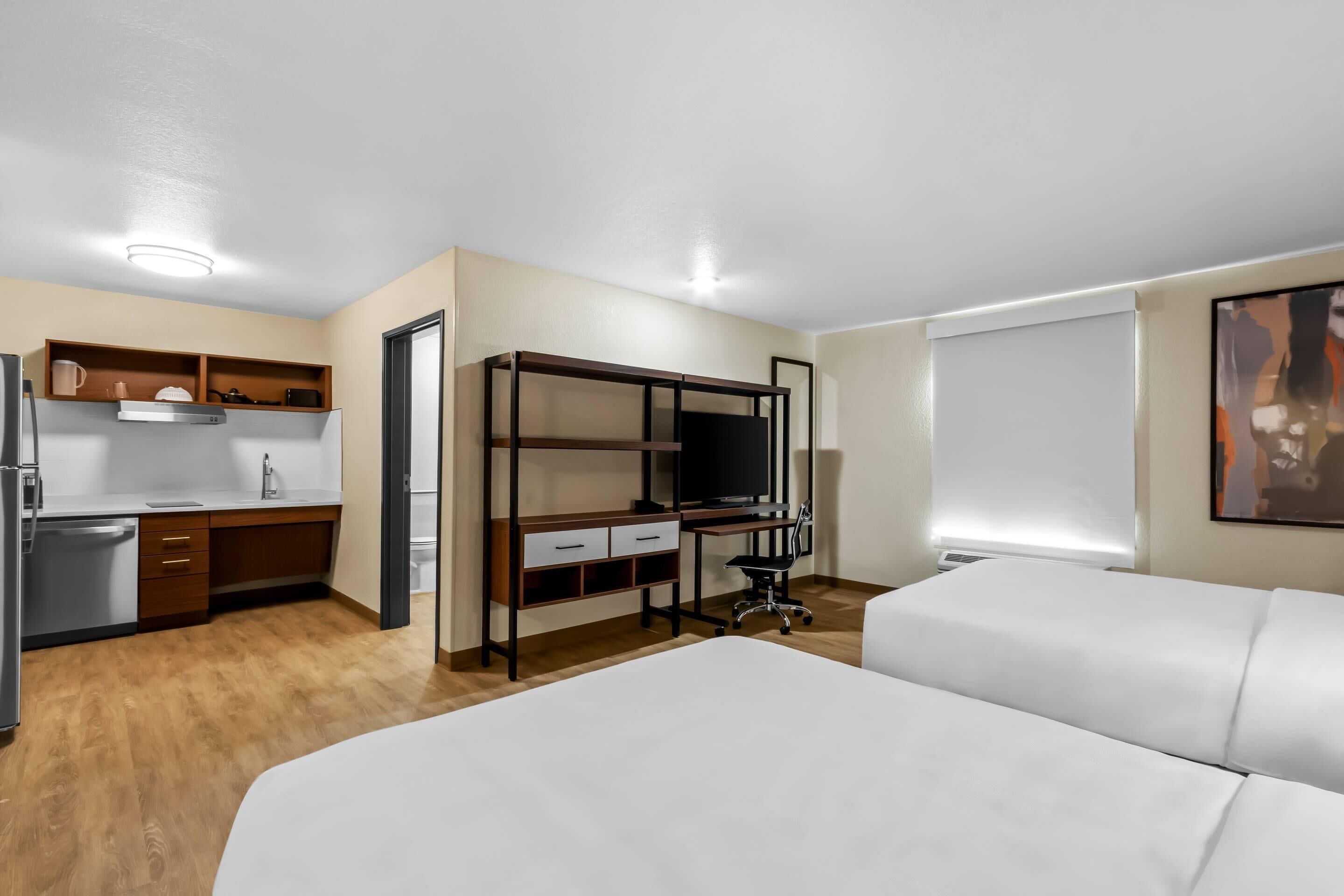 A hotel room with a bed, a desk with a computer, a chair, a small kitchenette, and a window.