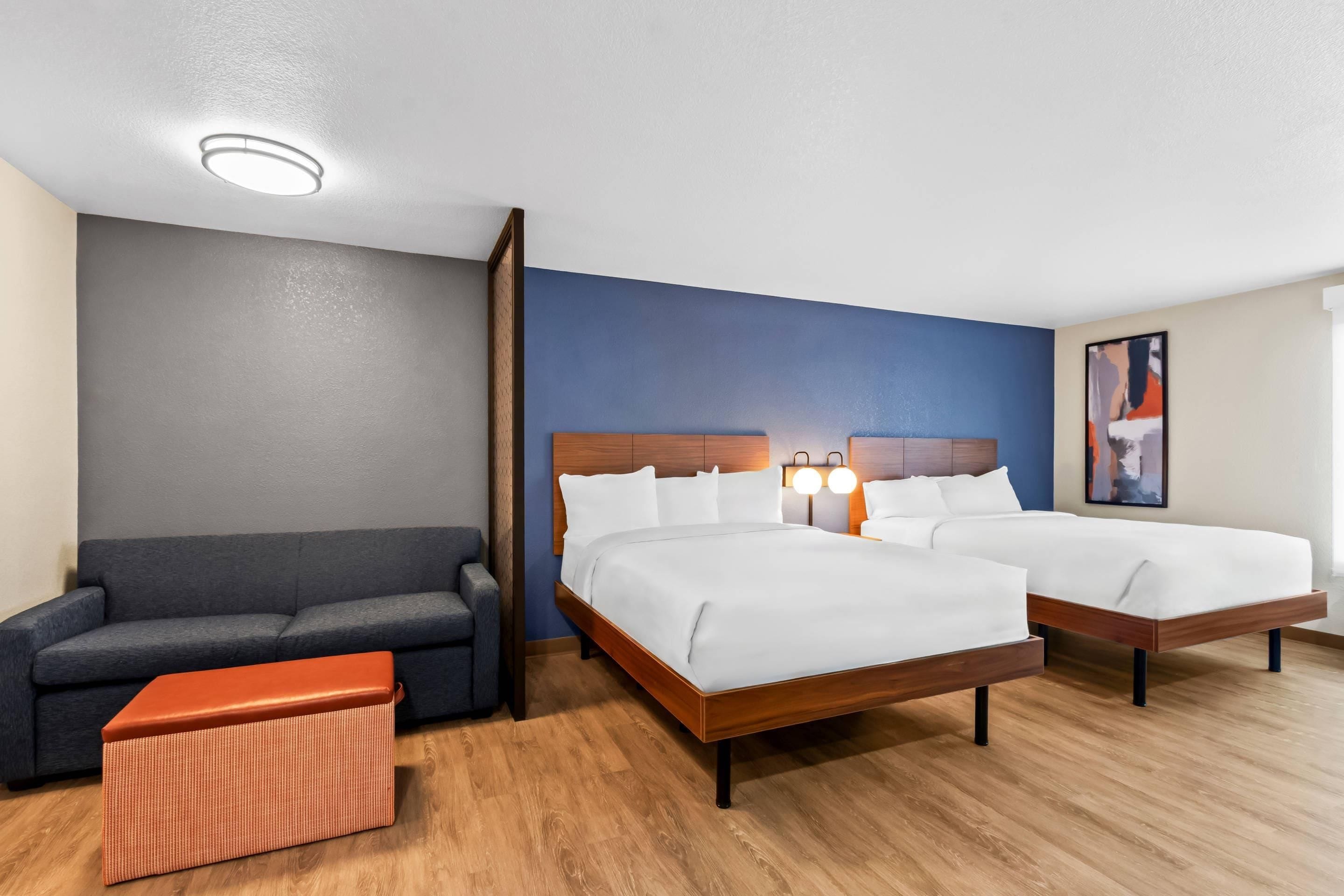 Une chambre d’hôtel avec deux lits, un canapé et un petit pouf orange.
