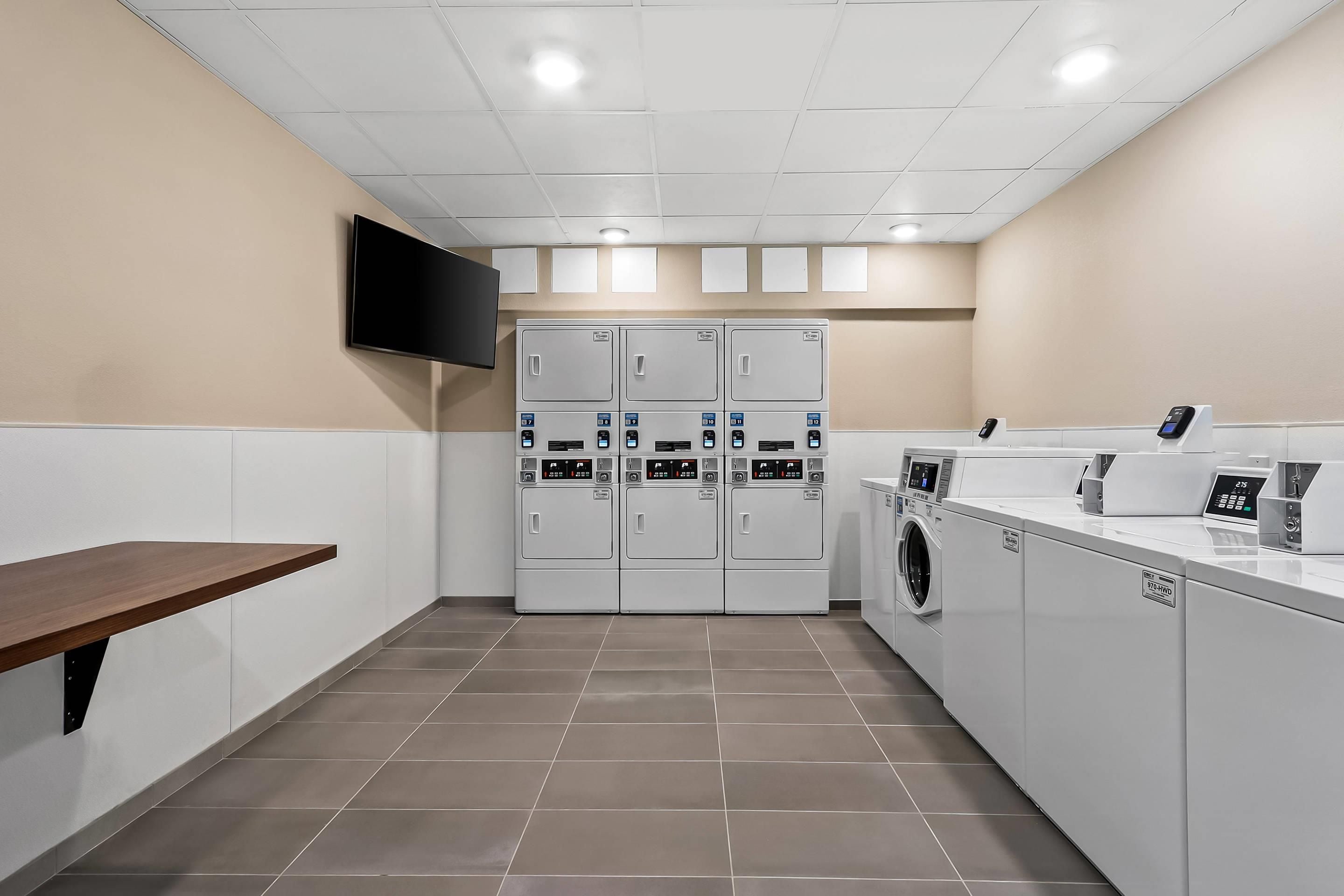 A laundry room with four stacked washers and dryers, a flat-screen TV mounted on the wall, and a wooden bench.