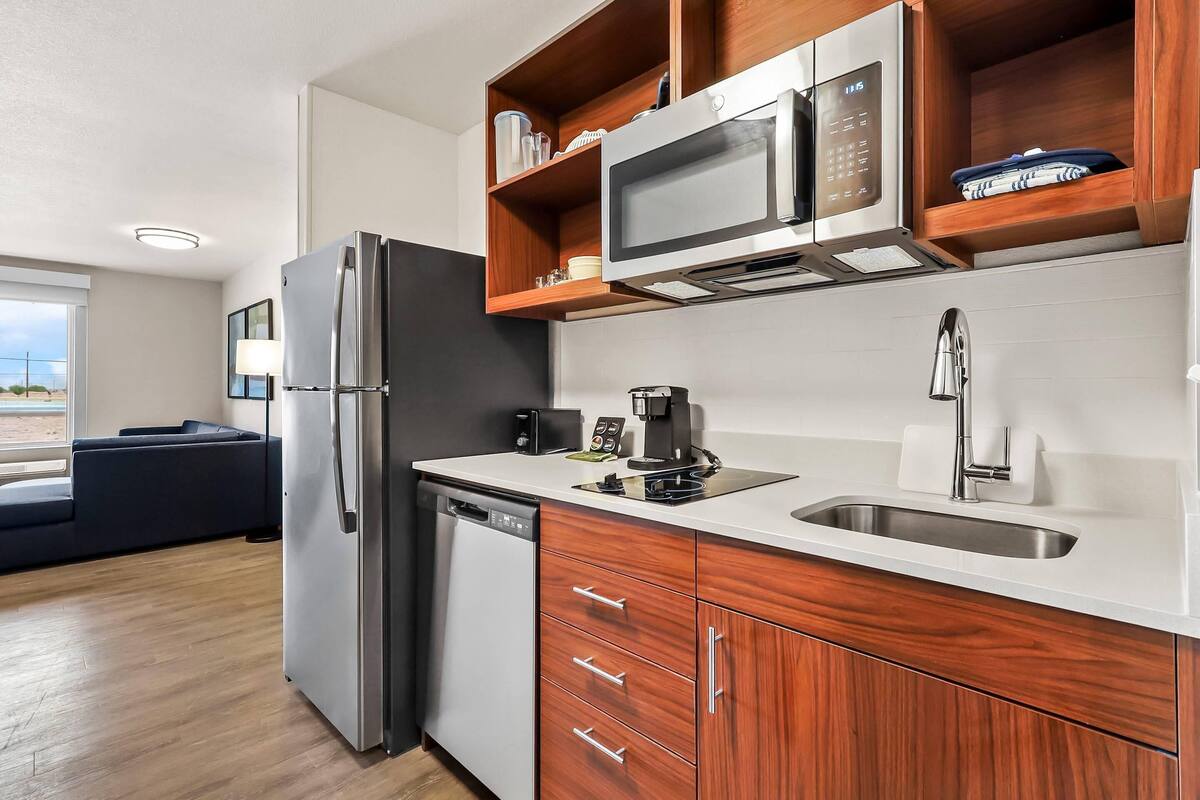 A modern kitchen with a refrigerator, microwave, and coffee maker. There is a wooden cabinet above the sink and a dishwasher to the left.