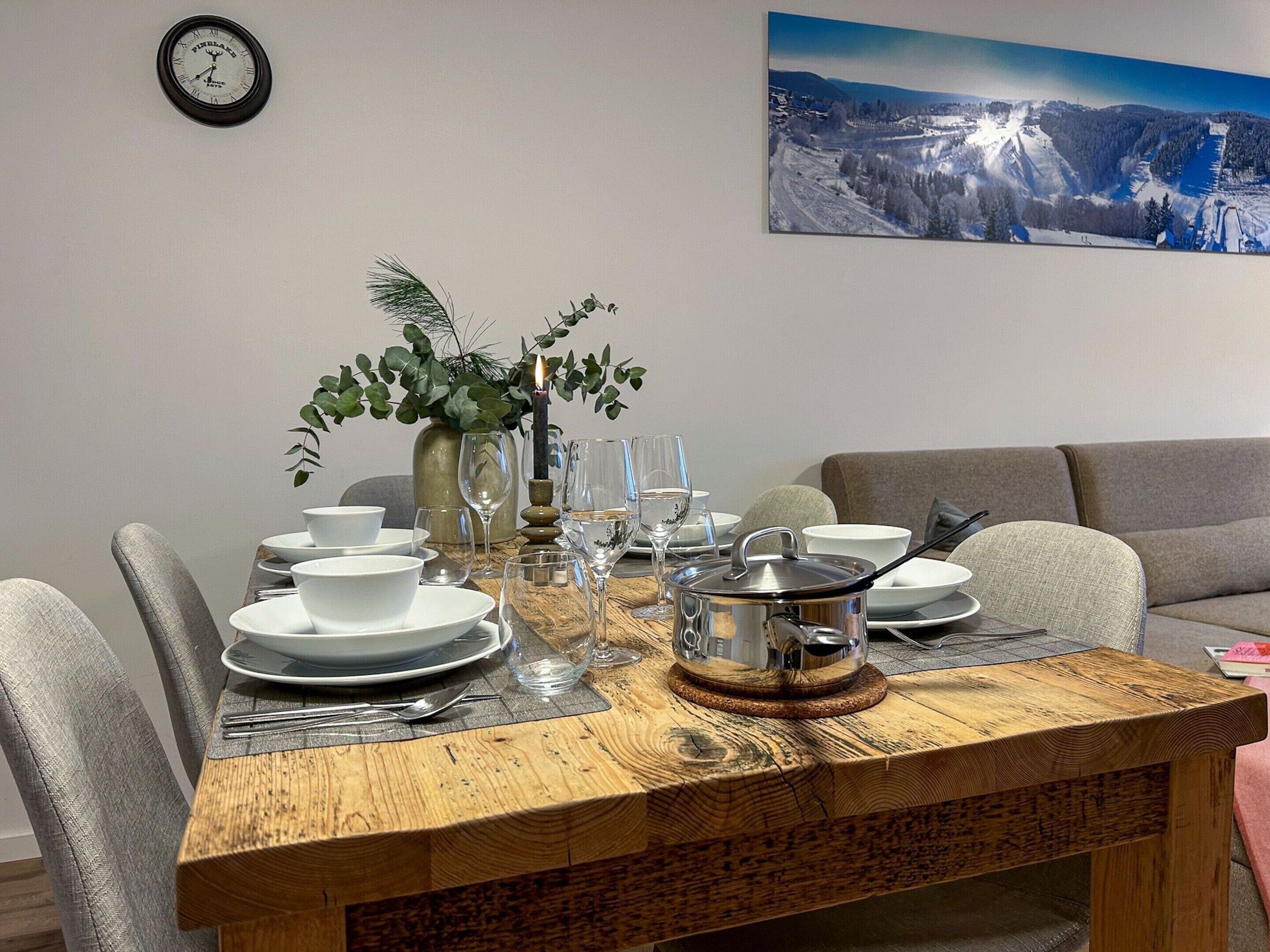 A dining table set with white plates, bowls, and cutlery, a vase with greenery, and a candle. A clock is on the wall, and a scenic painting is above the table.