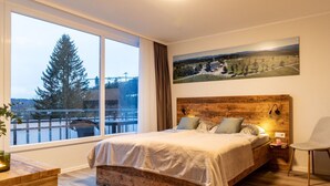 Une chambre avec une grande fenêtre, un lit, une chaise et une tête de lit en bois.