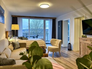 Une chambre d’hôtel comprenant un lit, un canapé, un fauteuil, une télévision et une grande fenêtre donnant sur des arbres, ainsi qu’un balcon.