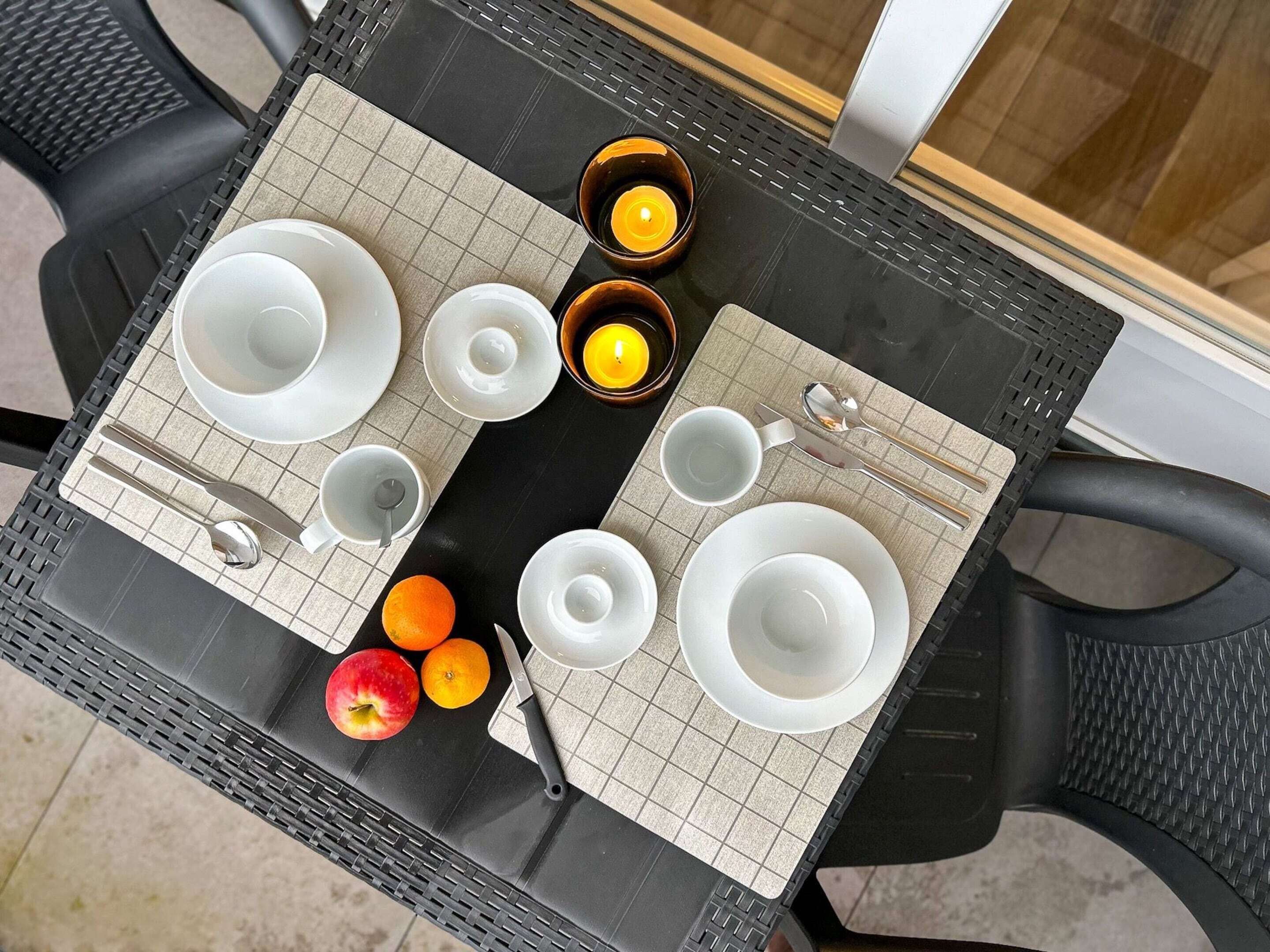 Una mesa dispuesta con vajilla blanca, cubiertos y dos velas encendidas, con frutas y un cuchillo sobre la mesa.