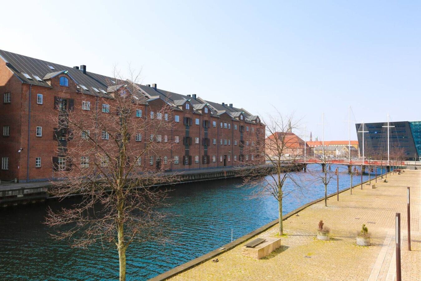 A riverside walkway with a brick building and a modern structure in the background.