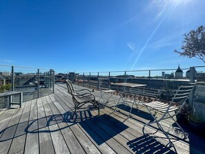 Una terraza de madera con sillas y mesas de metal, con vistas a un paisaje urbano de edificios y un cielo azul despejado.