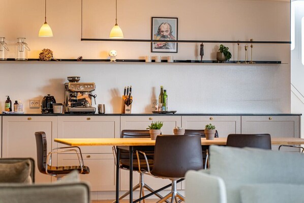 Una cocina moderna con una cafetera, estantes con diversos objetos y una zona de comedor con sillas y una mesa.